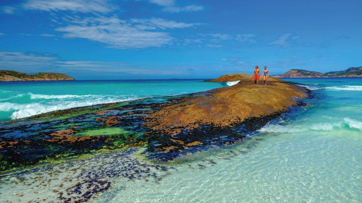 A couple walk along a submerged rock on a beach in Lucky Bay