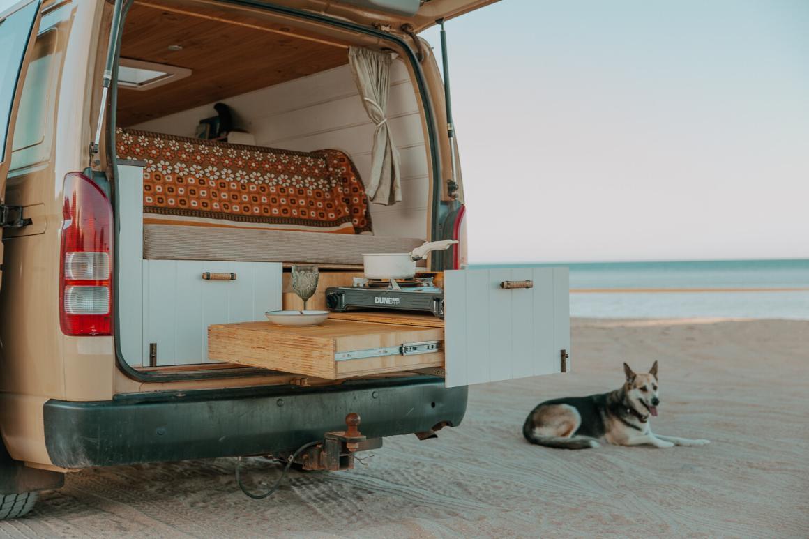 Marigold ‘Goldie’ The Hiace parked on a beach with the sea in the background. The back of the van is open with a kitchen set up and a dog relaxing on the beach. This is one of the best campervans to hire in Western Australia (WA).