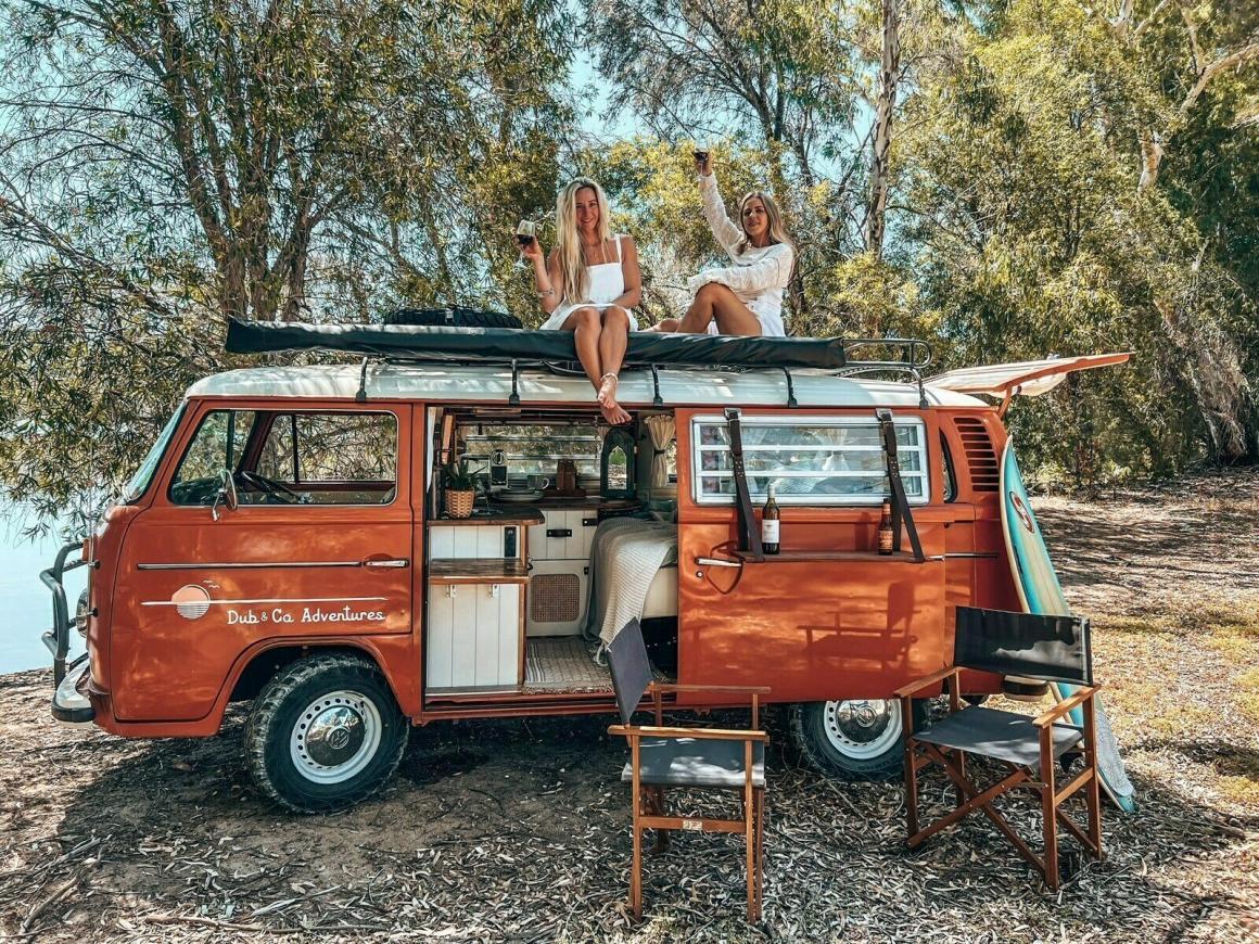 Two ladies sit on top of a Dub and Co campervan with wines in hand. This is one of the best campervans to hire in Western Australia (WA).