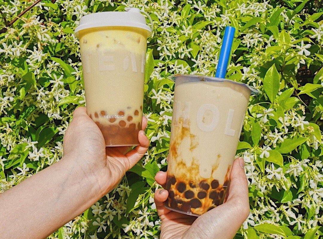 Two hands two teas with boba in front of a green bush. From Teaholic one of Perth's best bubble tea stores
