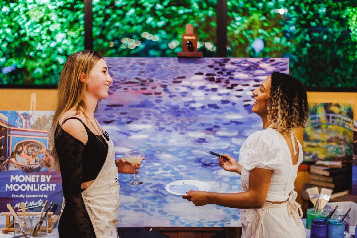 Two ladies stand in front of an easel with a painting on it, one with a paint tray in hand the other holding a cocktail as they paint at Monet in Paris