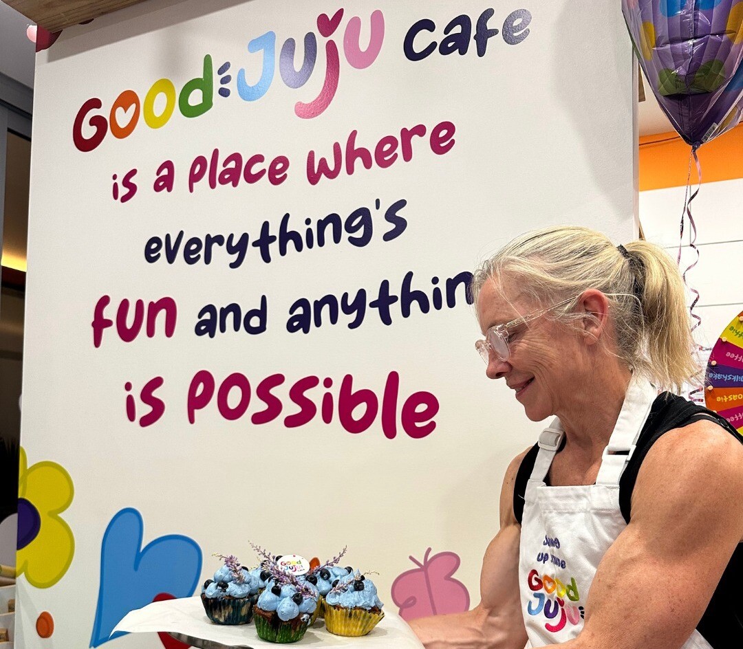 One of the owners of Good Juju Cafe holds up a tray of muffins in front of a sign in the Good Juju Cafe that reads "Good Juju Cafe is a place where everything's fun and anything is possible"