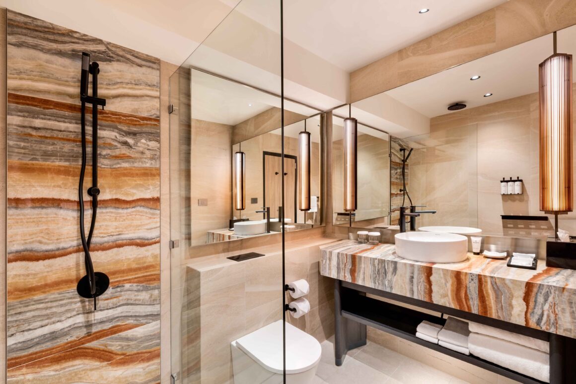 A wide angle photo of the bathroom in a Pan Pacific after it's refurb. Shows colourful stone wall shower, large mirror, funky long lights and a colourful stone sink. 