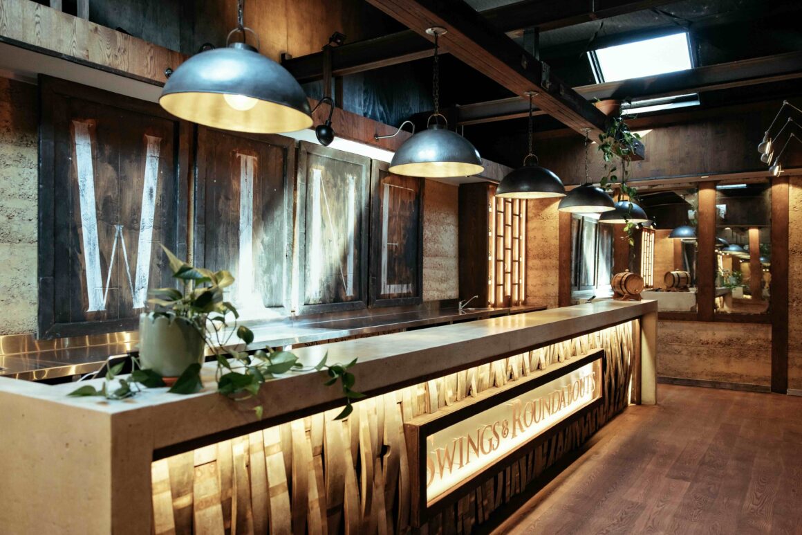 A wide angle view of the fully wooden tasting bar at the new cellar door of Swings & Roundabouts. Three large lighting fixtures are hung above the bar with the word "wine" printed on wood behind the bar.