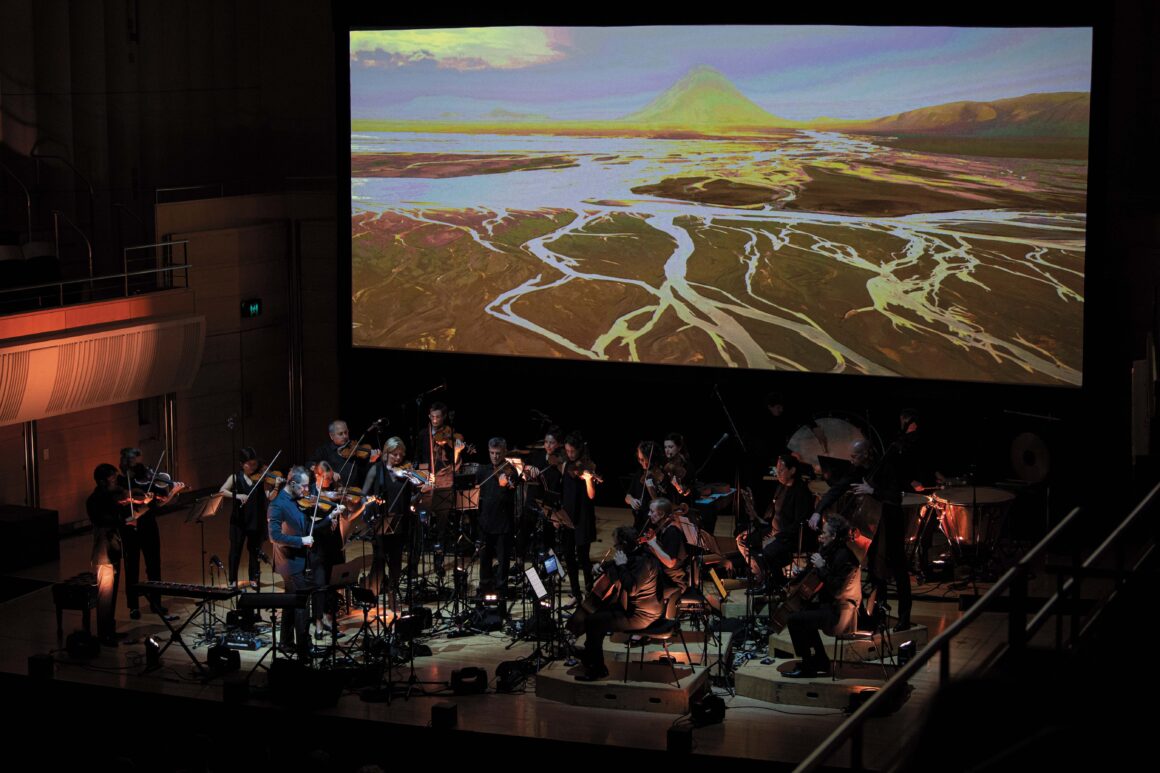 The Australian Chamber Orchestra performs on stage in front of a large screen playing thr River film