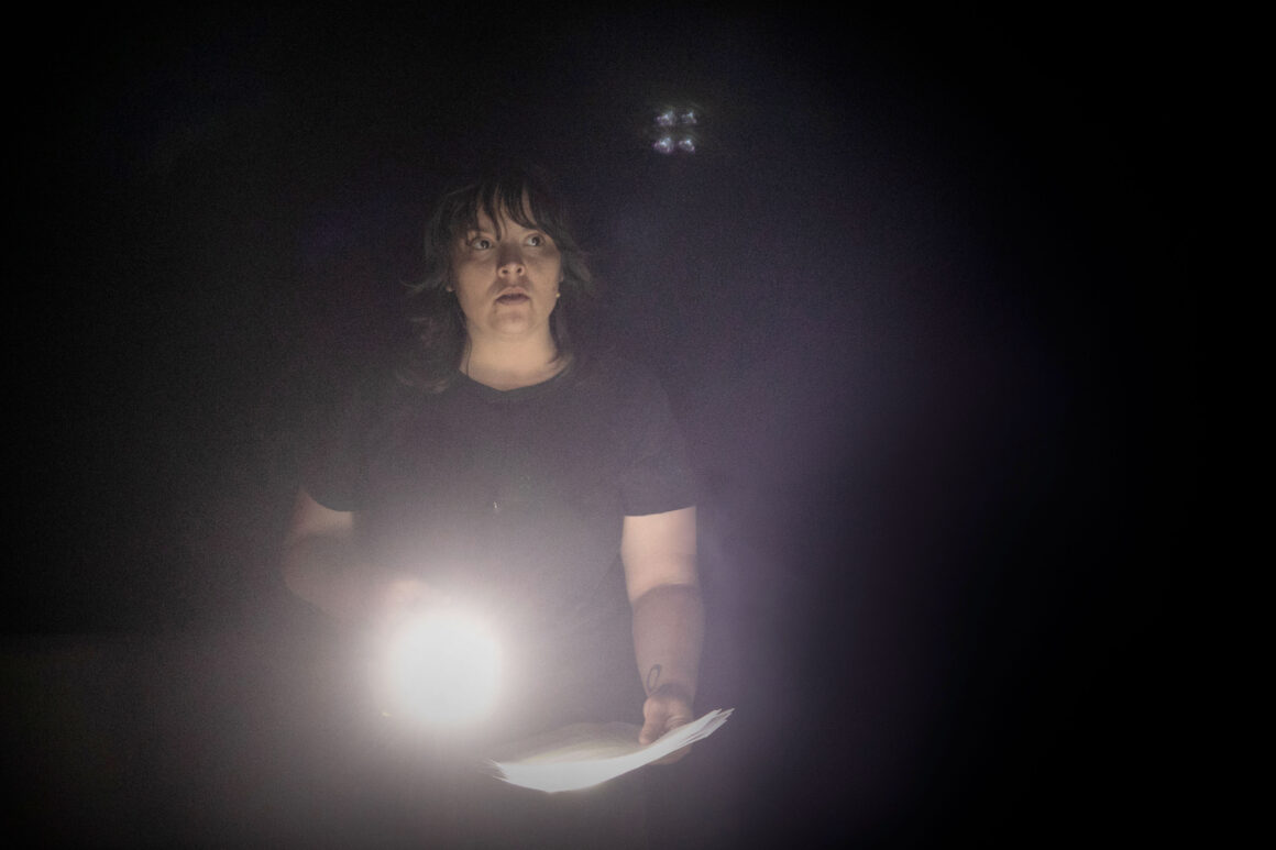 A woman stands in the dark with a paper in hand and torch in the other during the Logue Lake experience