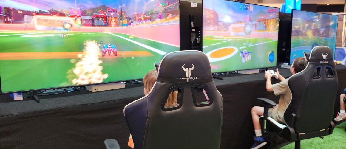 Two kids sitting in gaming chairs in front of screens, playing a video game