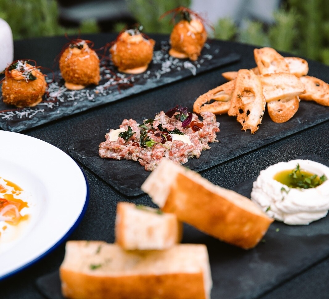 Lots of food dishes including arancini balls, pate with bread and hummus sit on a black table at Lygon Lane