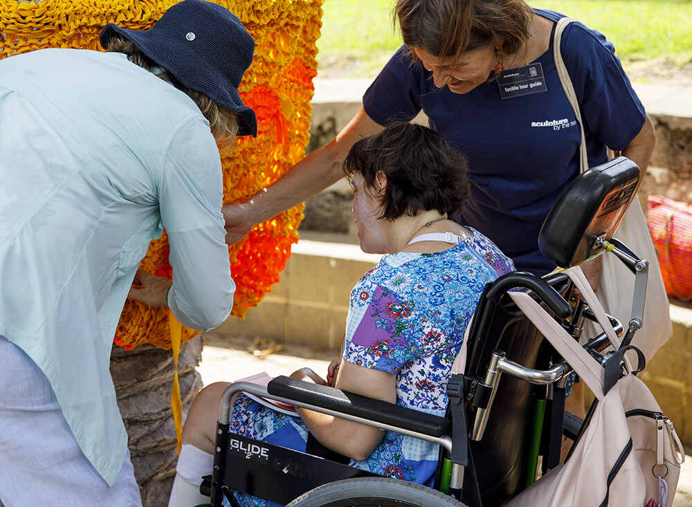 Two Sculpture by The Sea volunteers take a lady in a wheelchair on a tactile tour of the artwork