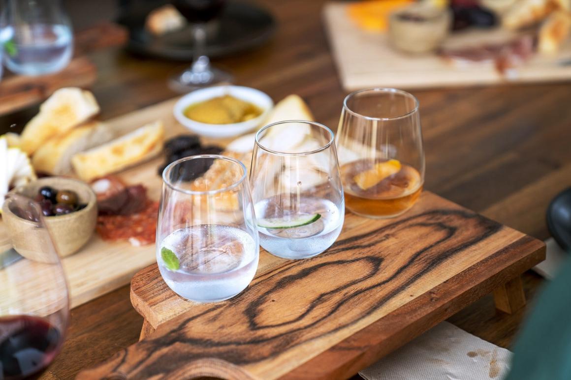 Three glasses with cocktails sit on a wooden board with a charcuterie behind it 