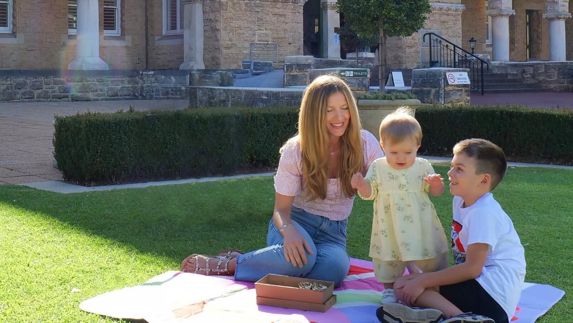 A mother sits with her daughter and son on a picnic rug on the lawns of The Perth Mint