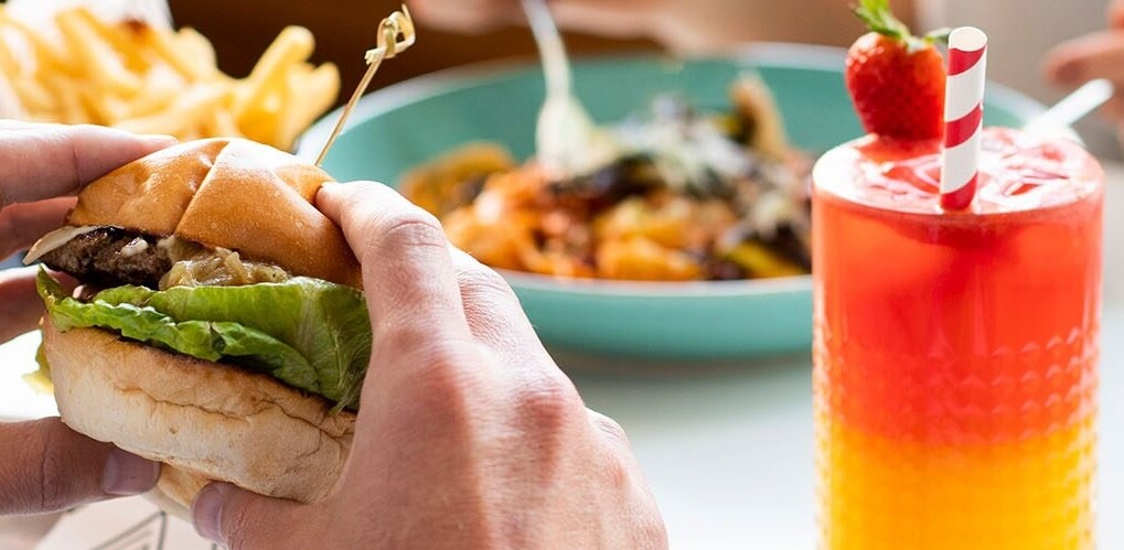 A man holds a burger on a table with sides and a drink. At Claremont during their spend and win promotion