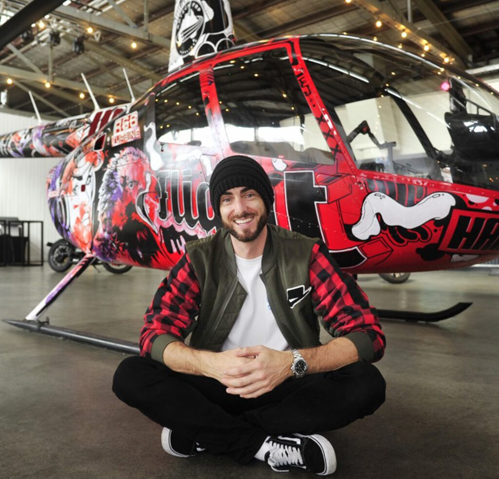 WA Artist Hans Bruechle (aka HANDBRAKE) sitting in front of his helicopter painted artwork. His newest exhibition Chaos Concludes will show at Fremantle's naval store in October.